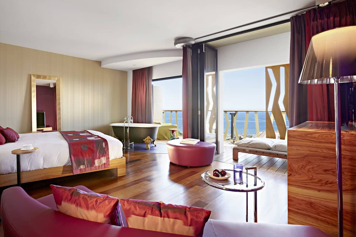 A modern hotel room with a large bed, a sitting area with a sofa and ottoman, a small round table with a glass, and a view of the sea through large windows.