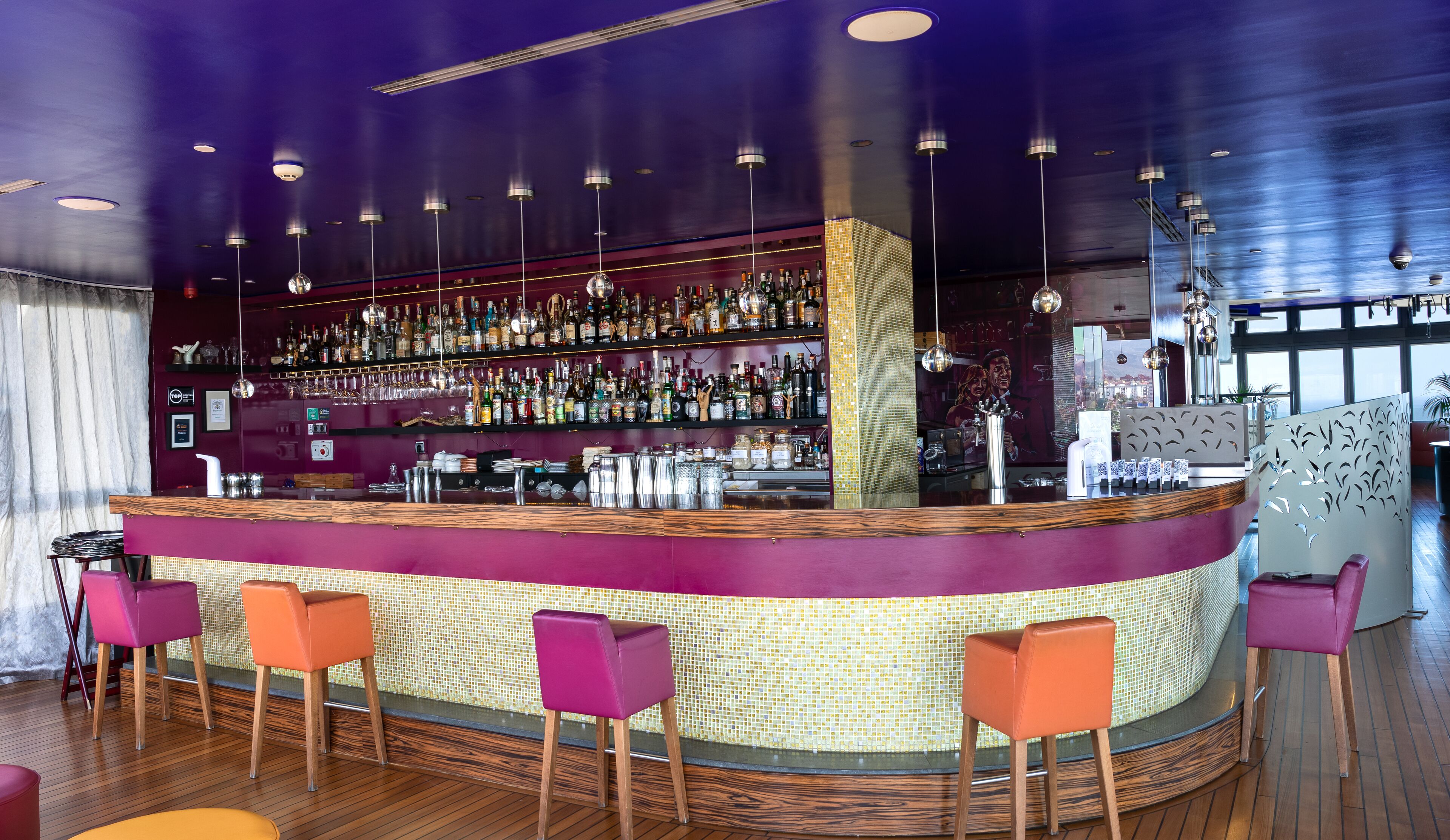 Eine Bar mit einer Holztresen, mehreren Flaschenregalen und bunten Stühlen.