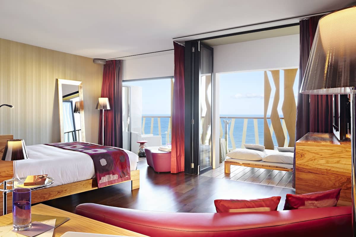 A modern hotel room with a large bed, a seating area, and a balcony with a view of the sea.