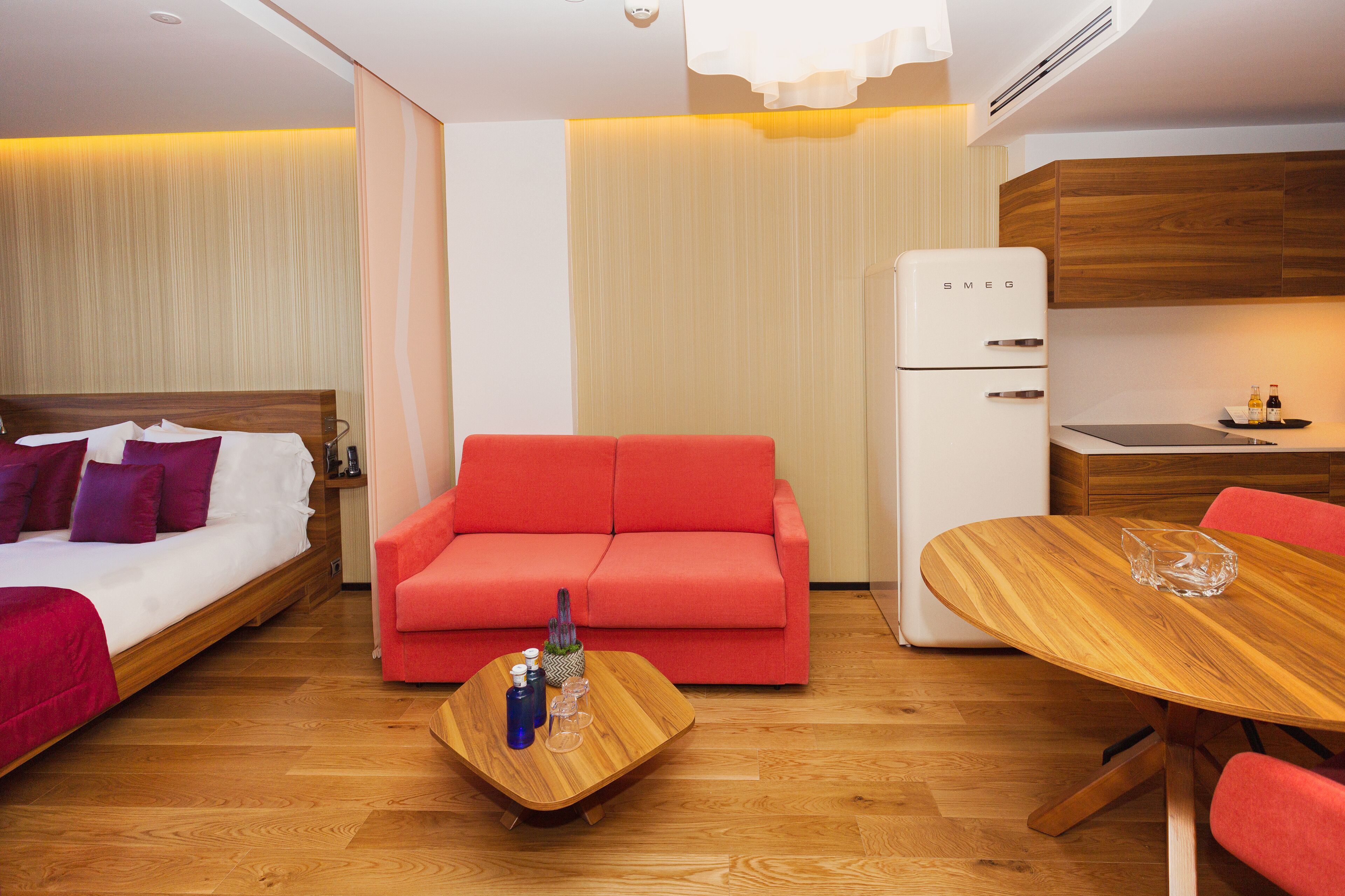 A modern hotel room with a bed, a red sofa, a wooden coffee table, a dining table, and a kitchenette.