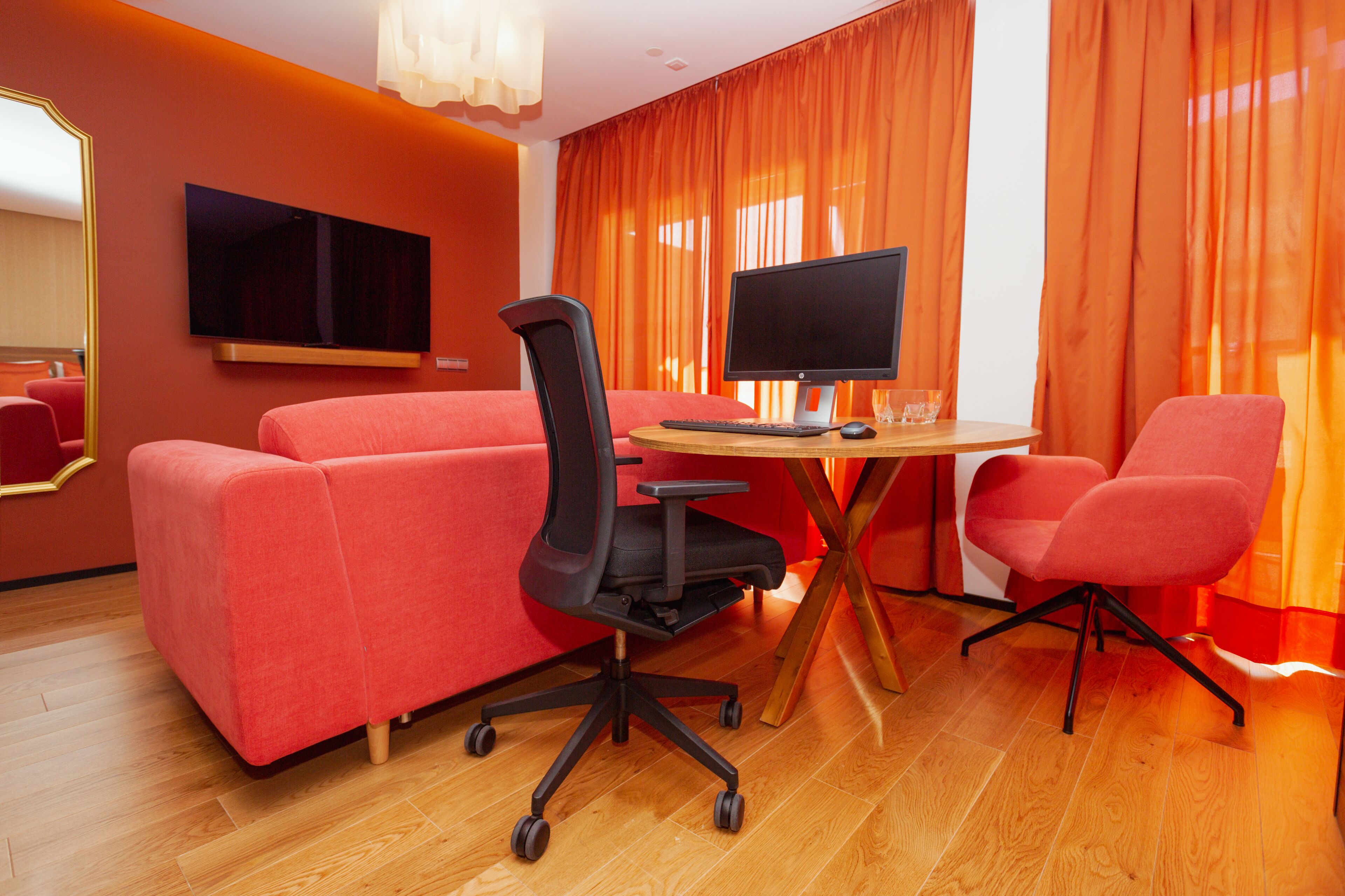 Una habitación de hotel moderna con un sofá rojo, una silla de oficina negra, una mesa de madera con una computadora y cortinas anaranjadas.