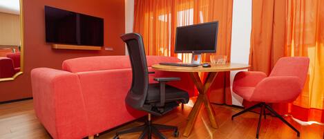 Una habitación de hotel moderna con un sofá rojo, una silla de oficina negra, una mesa de madera con una computadora y cortinas anaranjadas.