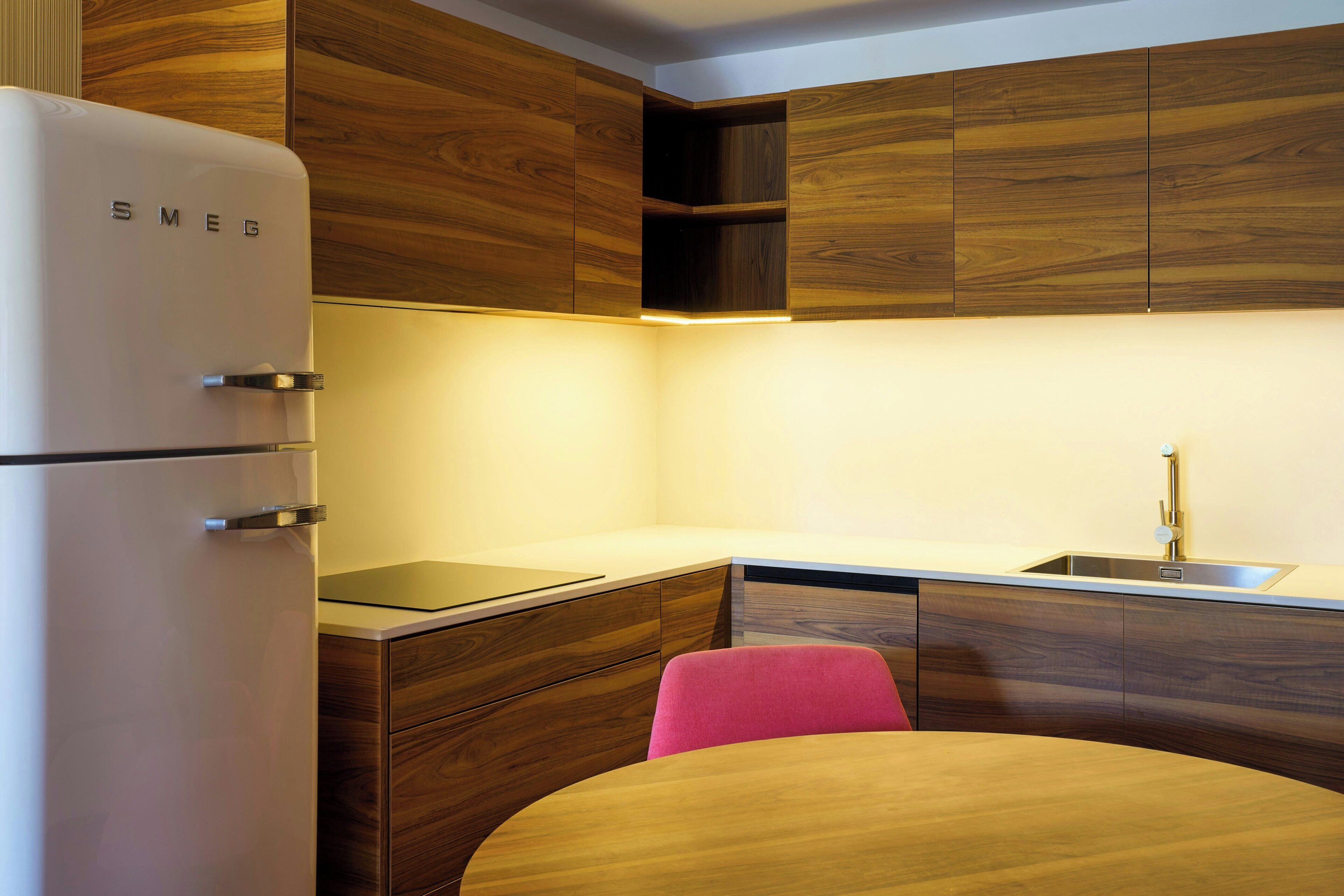 Una cocina compacta con un refrigerador blanco, armarios de madera, un lavamanos y una silla rosa.
