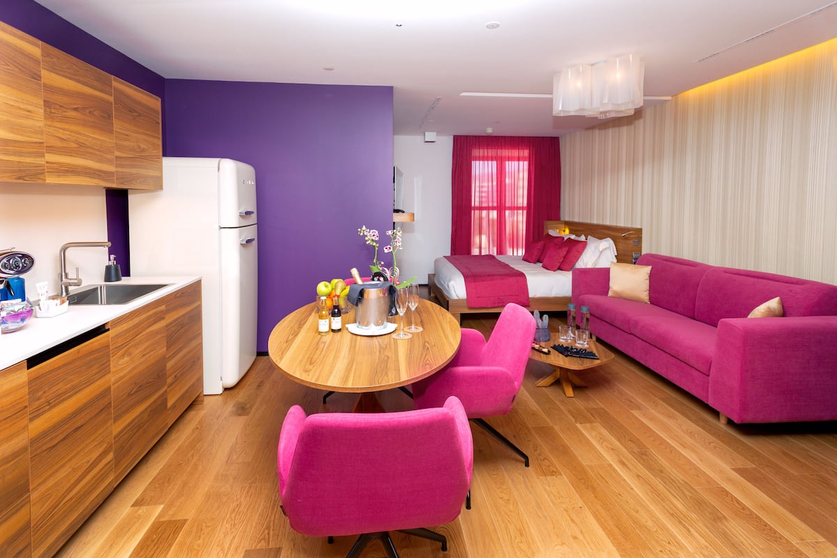 A modern living room with a pink sofa, a wooden dining table with chairs, and a kitchen area with a refrigerator and wooden cabinets.