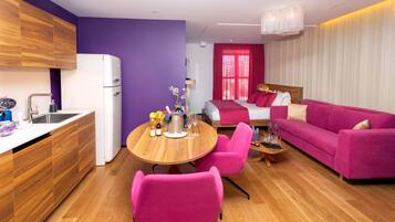 A modern living room with a pink sofa, a wooden dining table with chairs, and a kitchen area with a refrigerator and wooden cabinets.