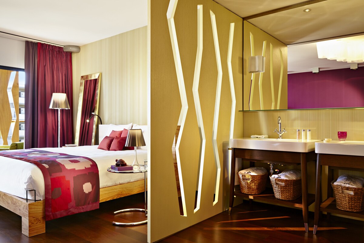 Habitación de hotel moderna con una cama grande, una mesita de noche, un espejo, un tocador con lavabo e iluminación decorativa en la pared.