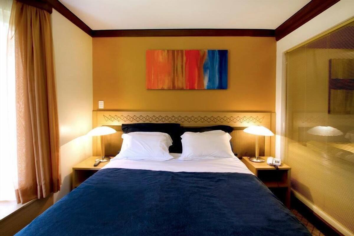 A hotel room with a bed, two bedside lamps, a painting on the wall, and a window with blinds.