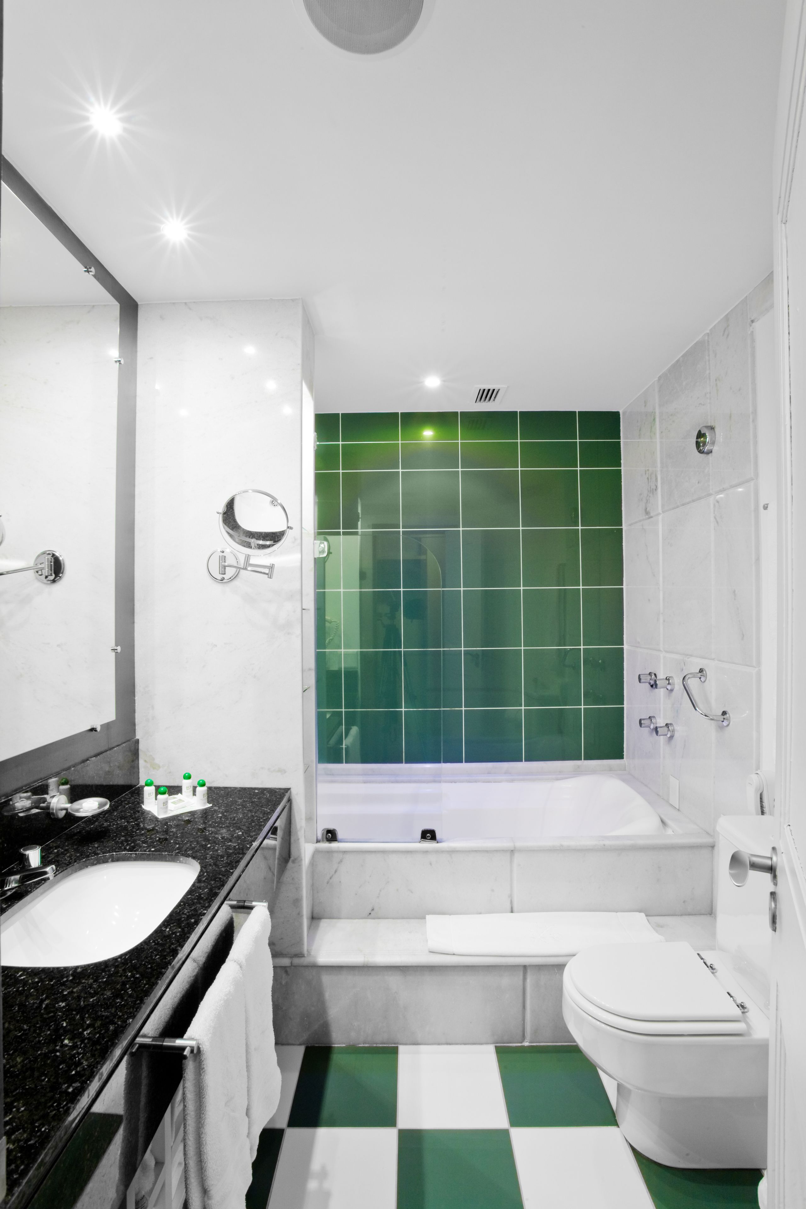 A modern bathroom with a white sink, mirror, and toilet. It features a green-tiled shower wall and a checkered floor.