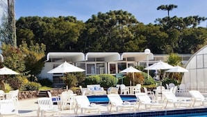 Zona de piscina con sillones blancos y sombrillas, rodeada de vegetación y arquitectura moderna.