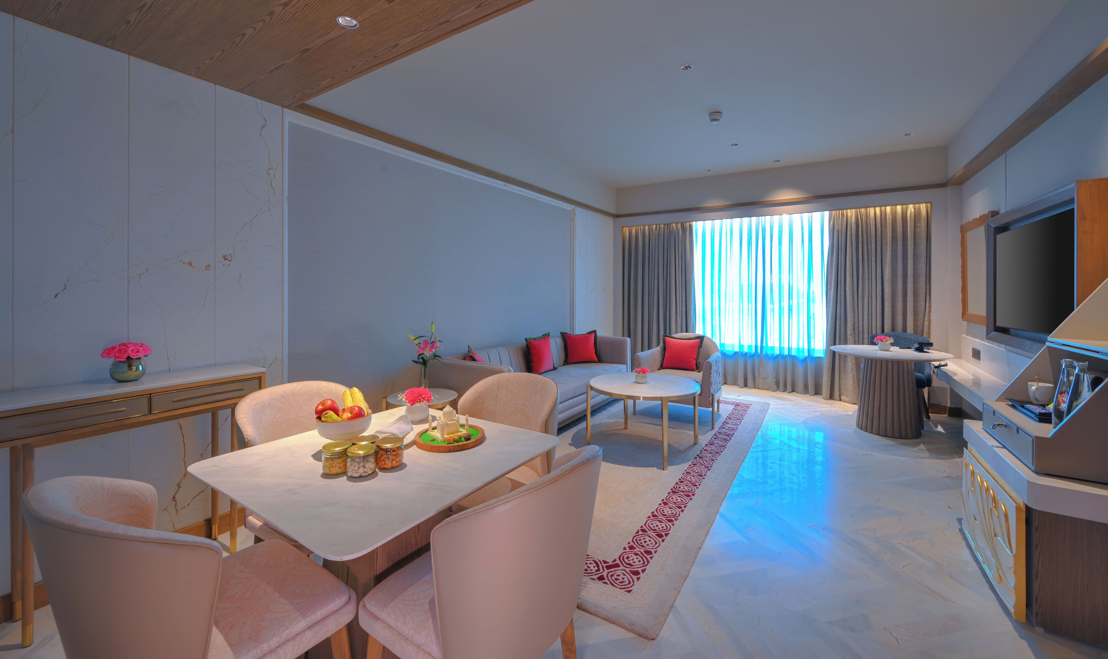 Una moderna camera d'albergo con zona pranzo, divano con cuscini rossi, televisore e un'ampia finestra con tende.