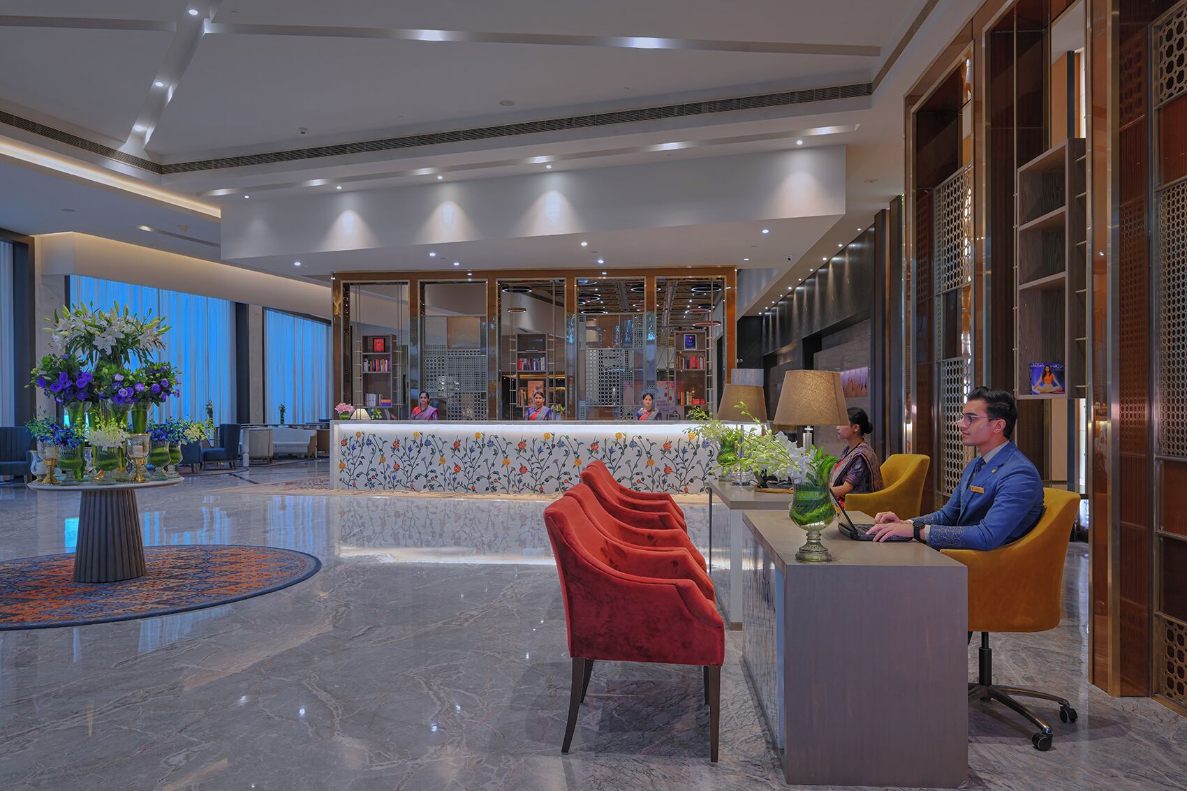 A hotel lobby with a reception desk, a red chair, and a man sitting at a table.
