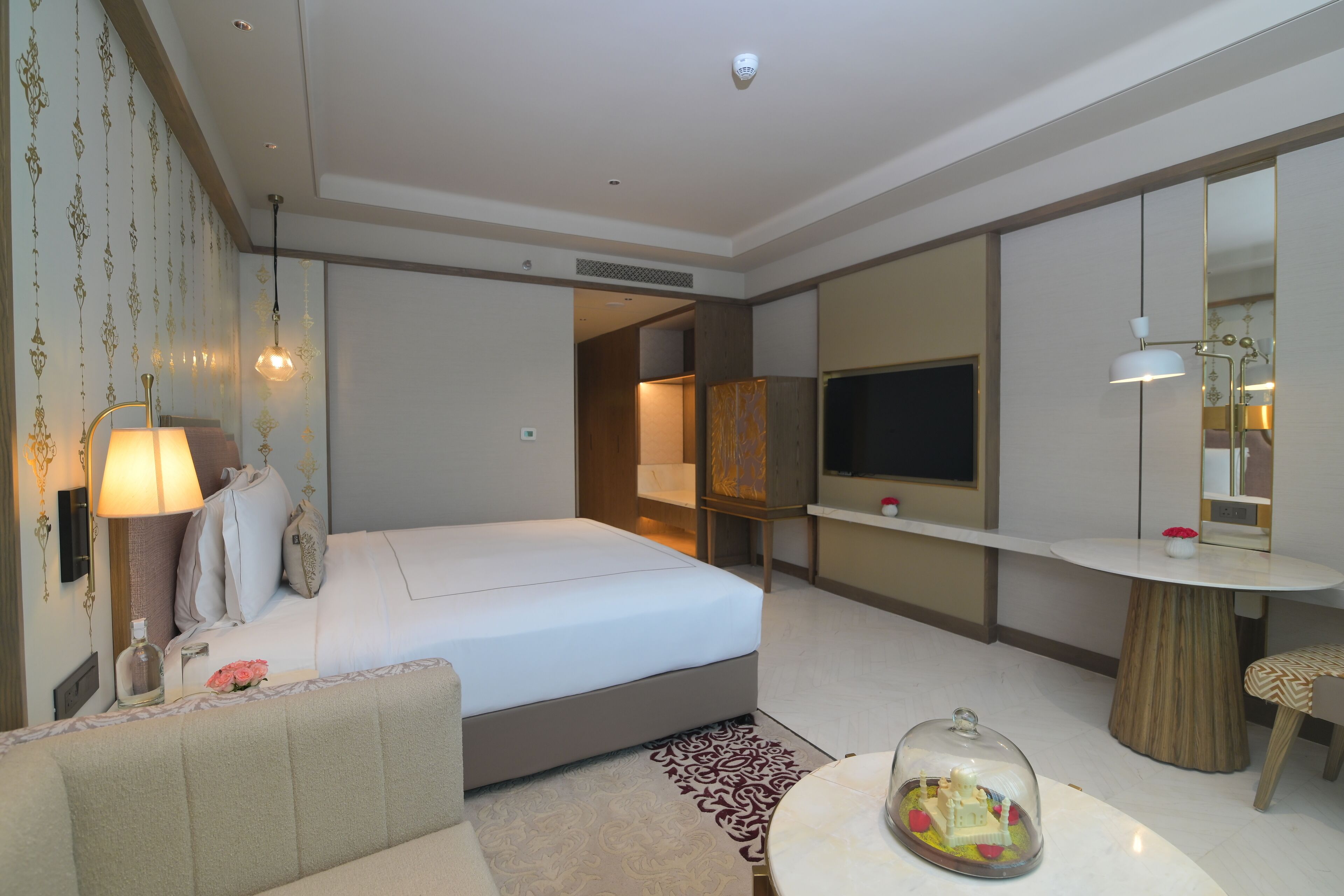 Una habitación de hotel con una cama, una mesita de noche con lámpara, un televisor, un espejo y una pequeña mesa redonda con un objeto decorativo.