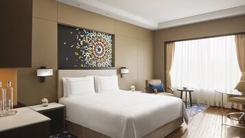 A hotel room with a large bed, a desk with a lamp, a chair, a small table with a plant, and a window with curtains.