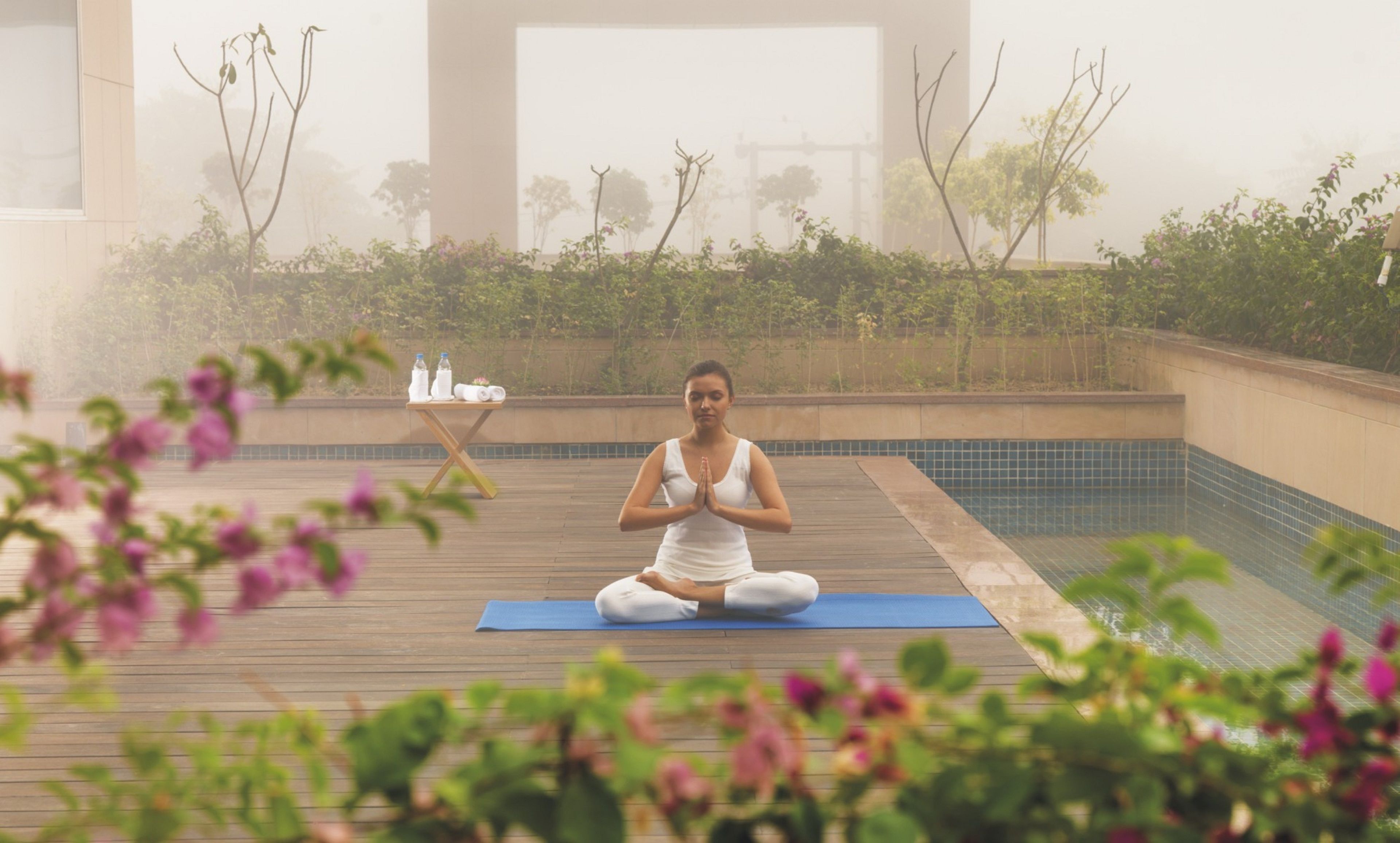 A person meditating on a wooden deck with a blue mat, overlooking a swimming pool.