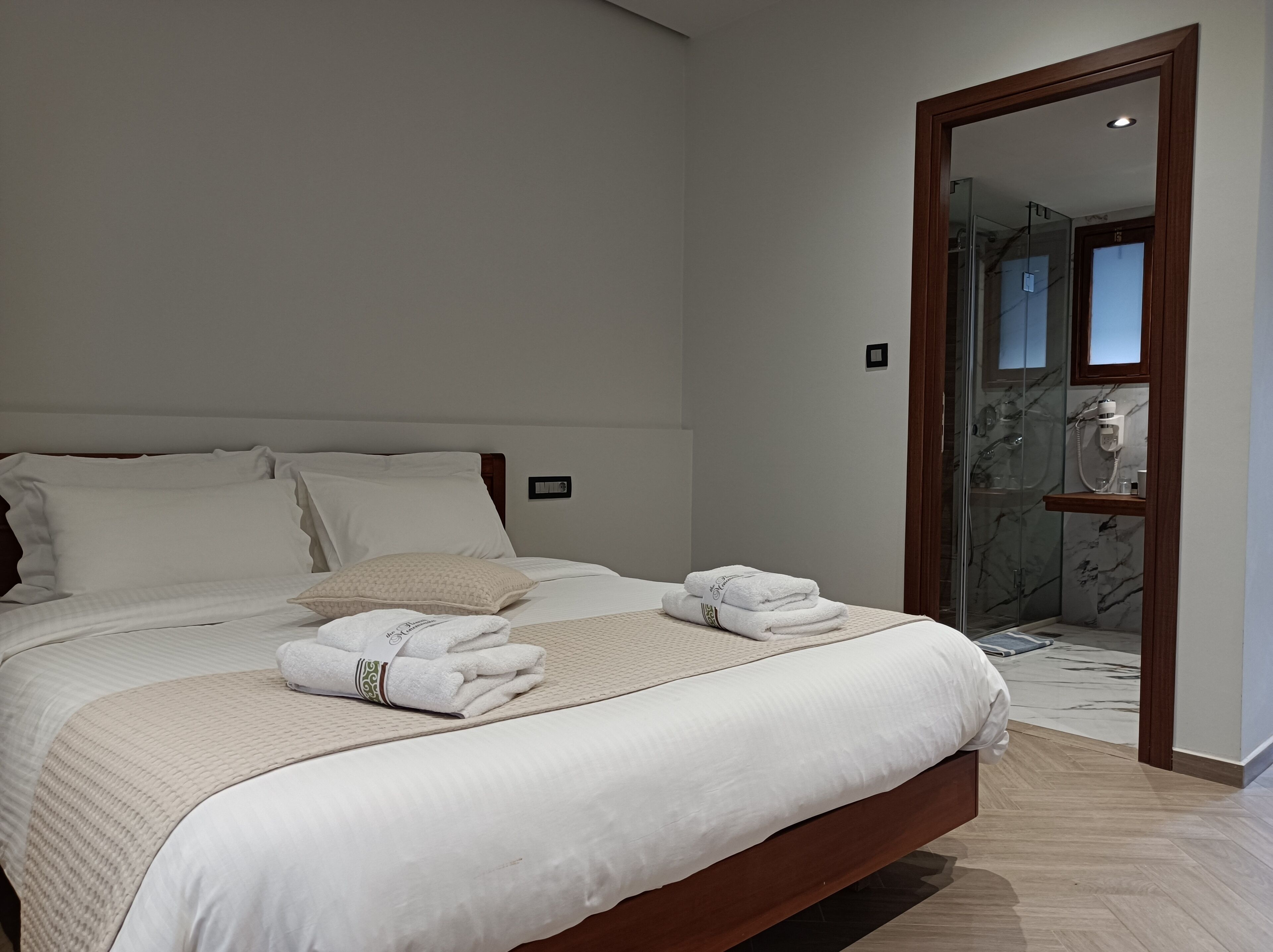 A neatly made bed with white linens and a beige throw blanket, a wooden headboard, and a small wooden shelf with toiletries.