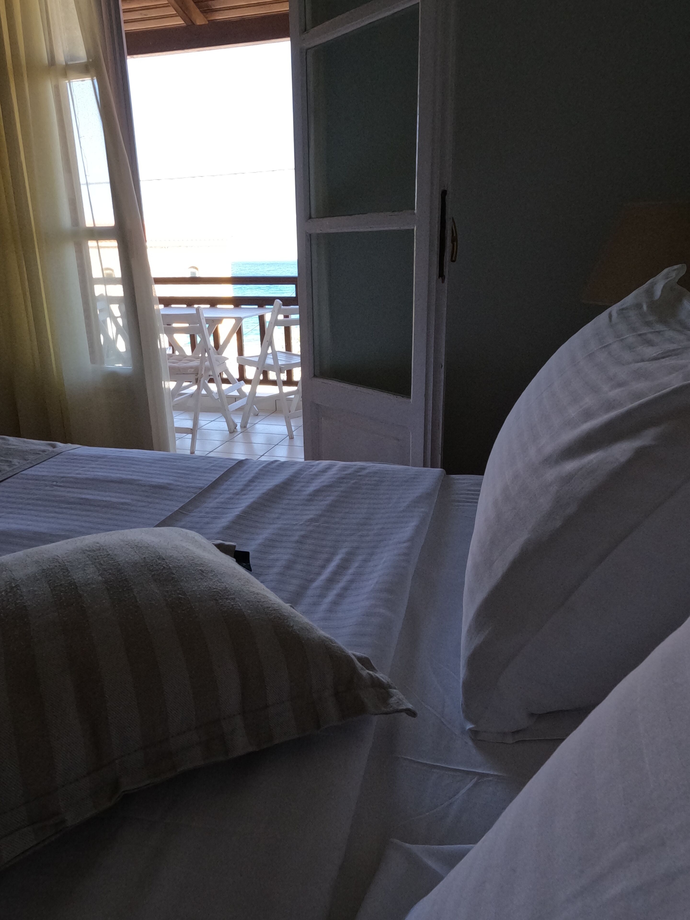 A bed with white linens and striped pillows, a wooden headboard, and a view of an outdoor deck with white chairs.