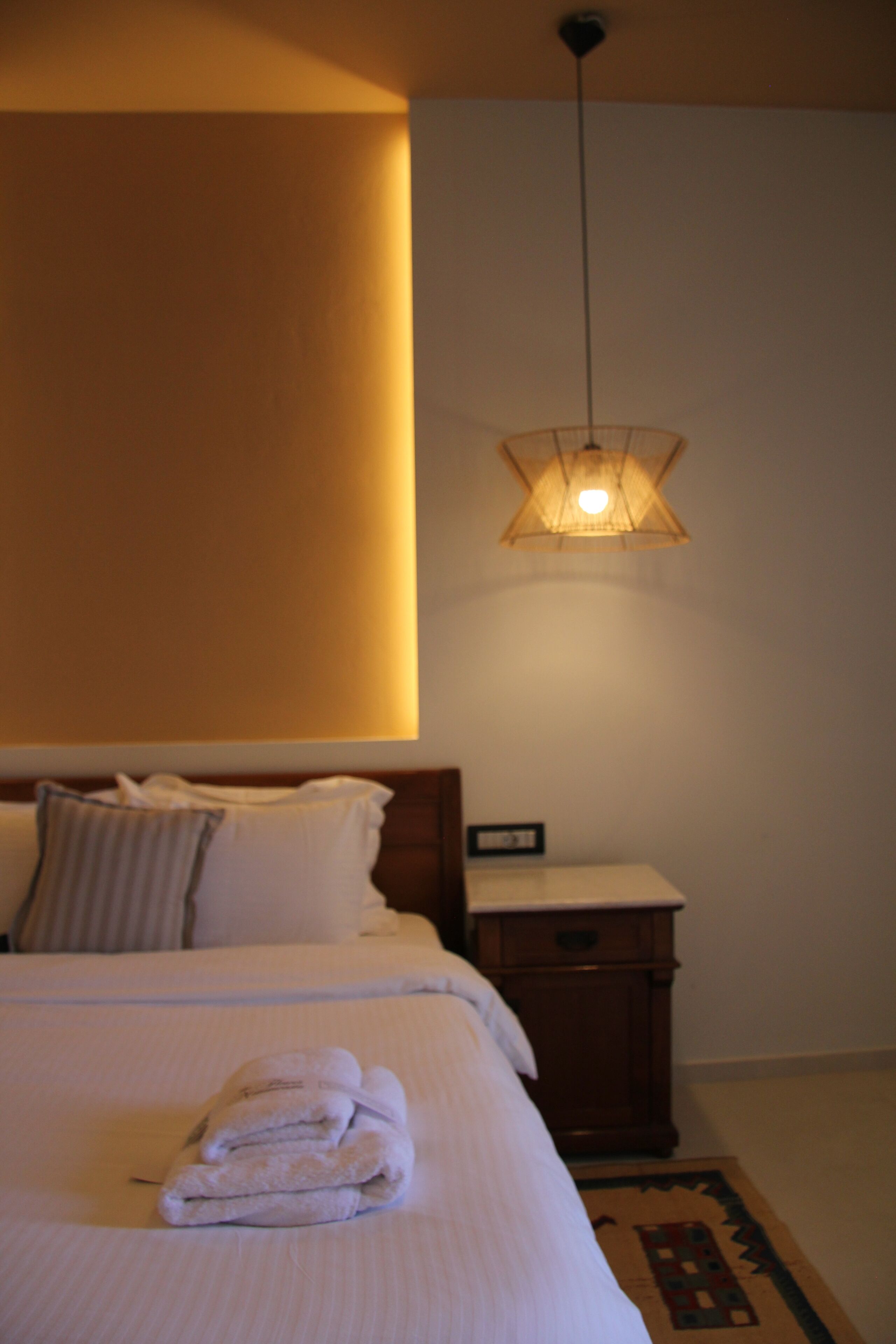 A neatly made bed with a folded towel, a bedside table with a clock, and a hanging light fixture.