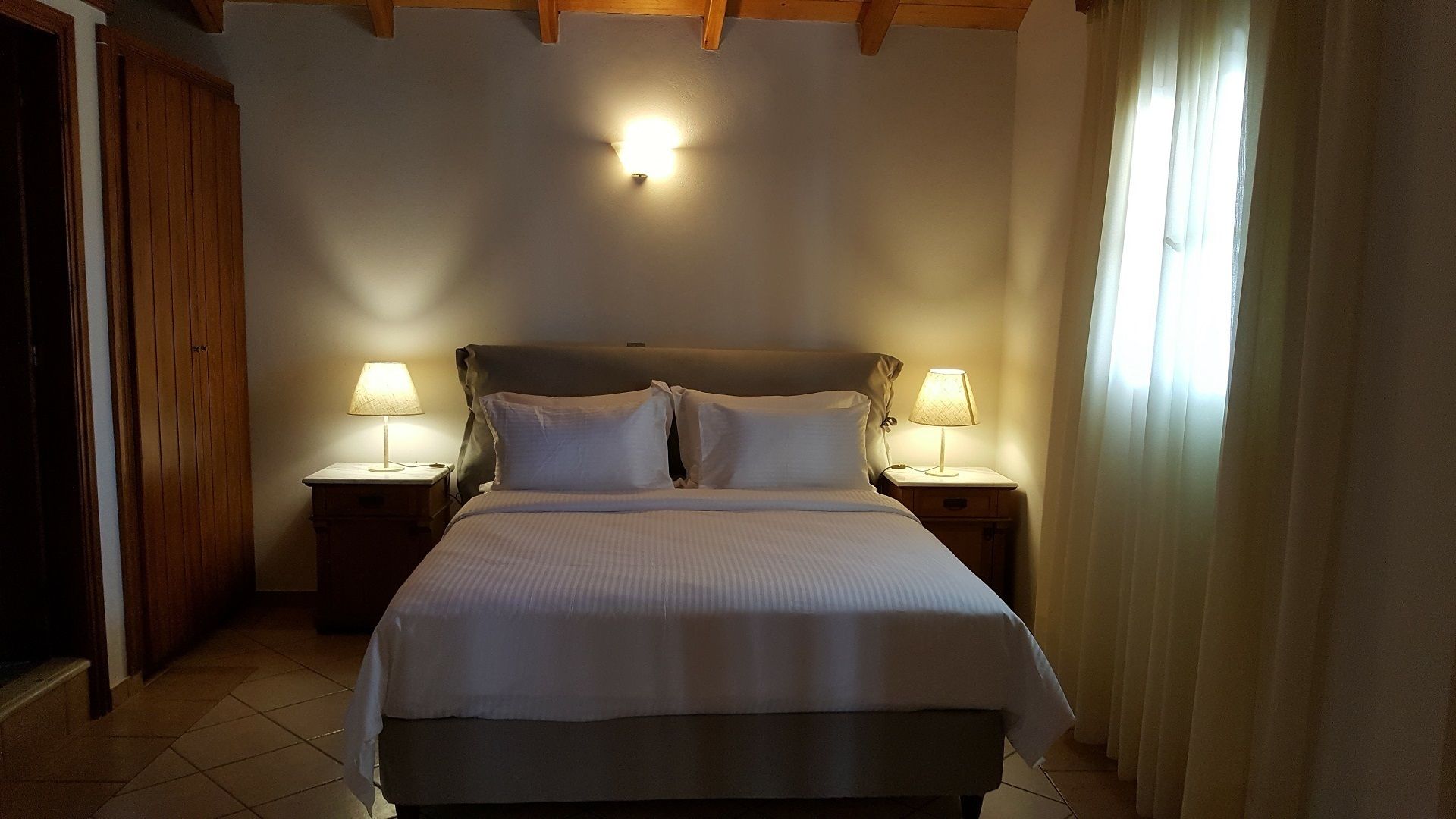 A bedroom with a bed, two bedside tables with lamps, wooden ceiling beams, and a window with curtains.