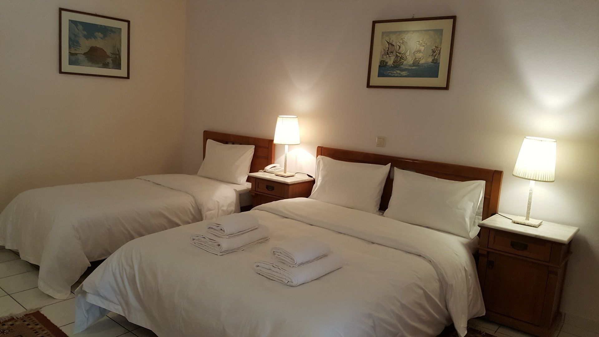 Ein Hotelzimmer mit zwei Betten, jeweils mit einem Holzkopfstück, weißen Laken und Nachttischlampen. An den Wänden befinden sich zwei Gemälde.
