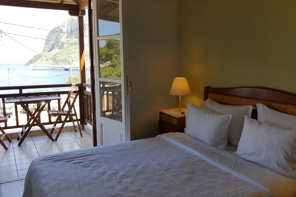 A bedroom with a bed, a nightstand, a lamp, and a view of the sea through an open door.