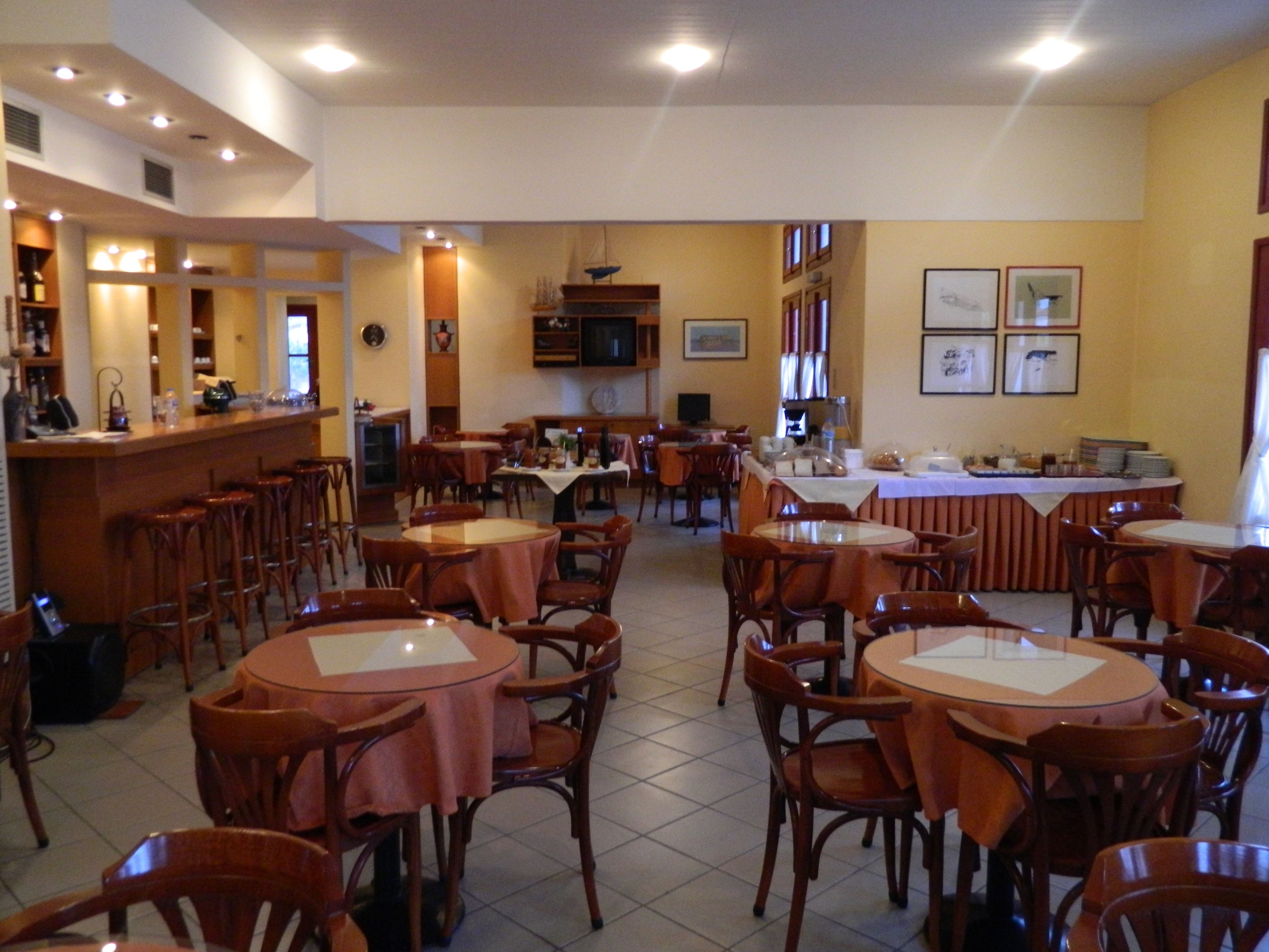 Un restaurante con varias mesas y sillas redondas, una zona de bar con taburetes y un televisor montado en la pared.