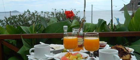 A table set with breakfast items including fruit, juice, and a slice of cake, overlooking a coastal view.