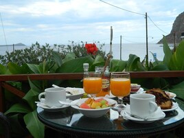 Una mesa dispuesta con alimentos para el desayuno, como fruta, jugo y una rebanada de pastel, con vista al mar.