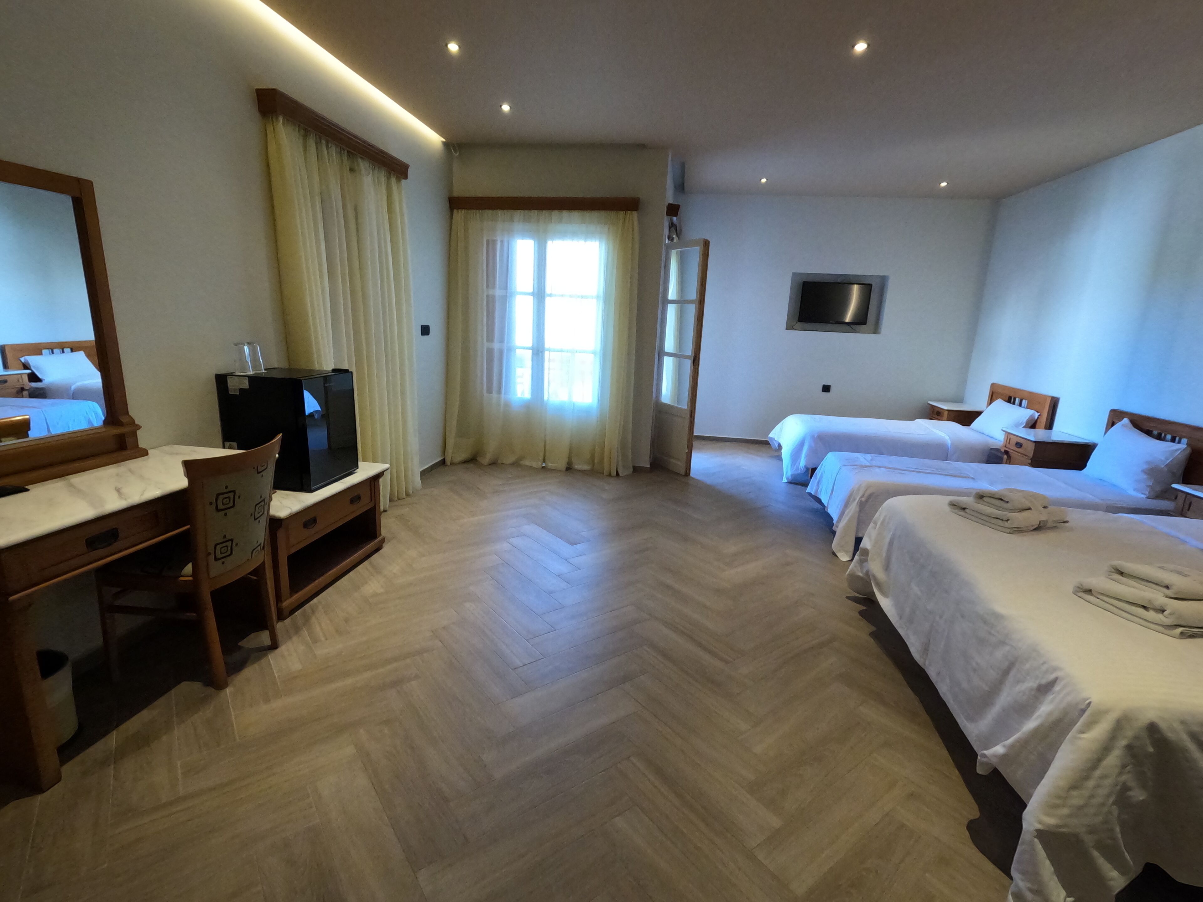 A hotel room with a wooden desk, a television, two beds, and a large window with curtains.