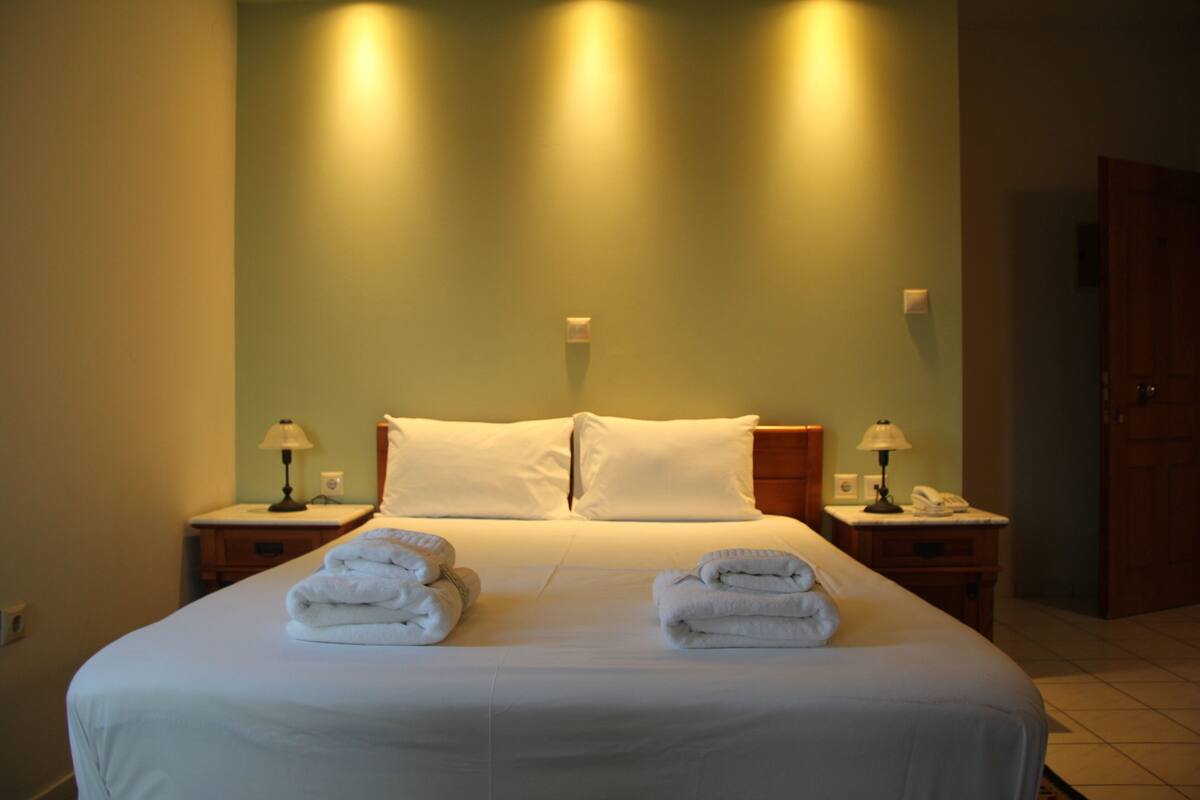 A neatly made bed with white linens and two folded towels on the bed. There are two bedside tables with lamps and a telephone. The room has a wooden headboard and a green wall.