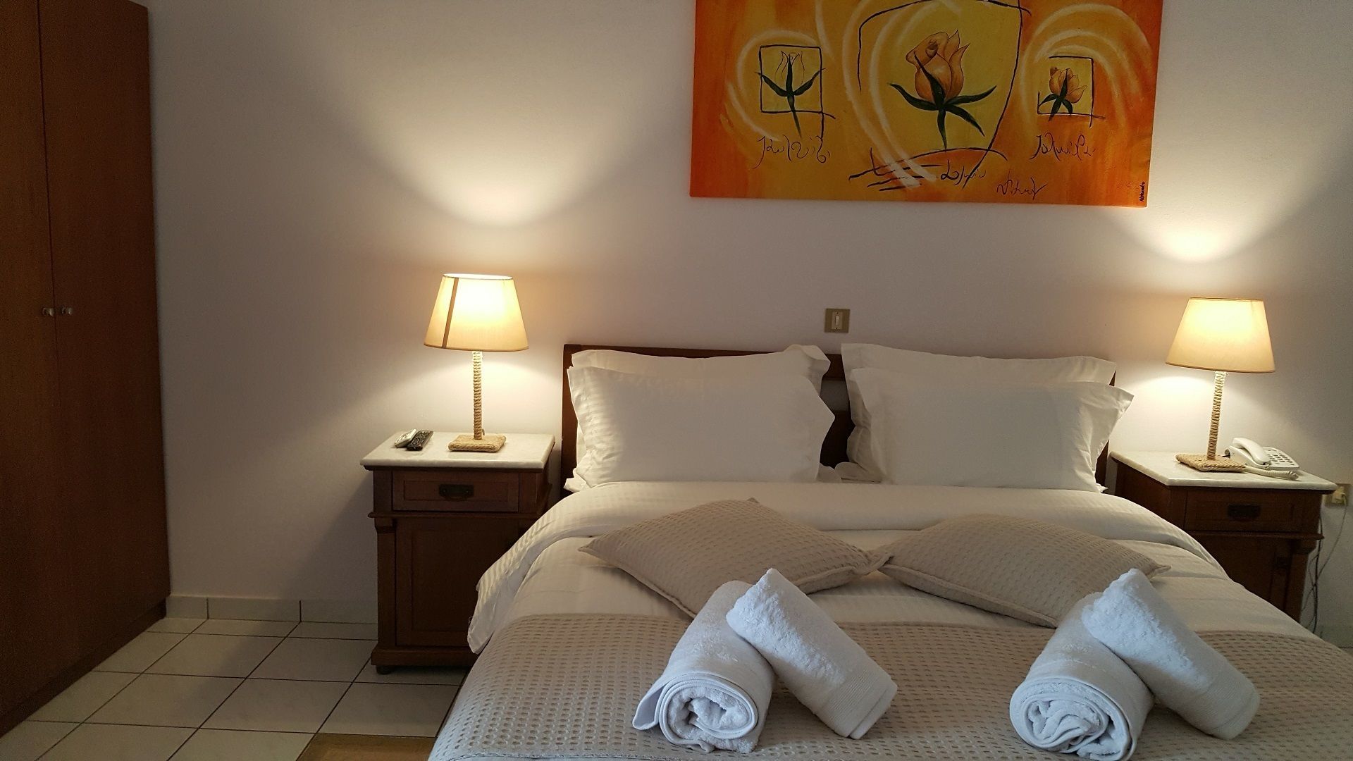 A neatly made bed with white linens and two rolled towels on the bed. There are two bedside tables with lamps and a painting above the bed.