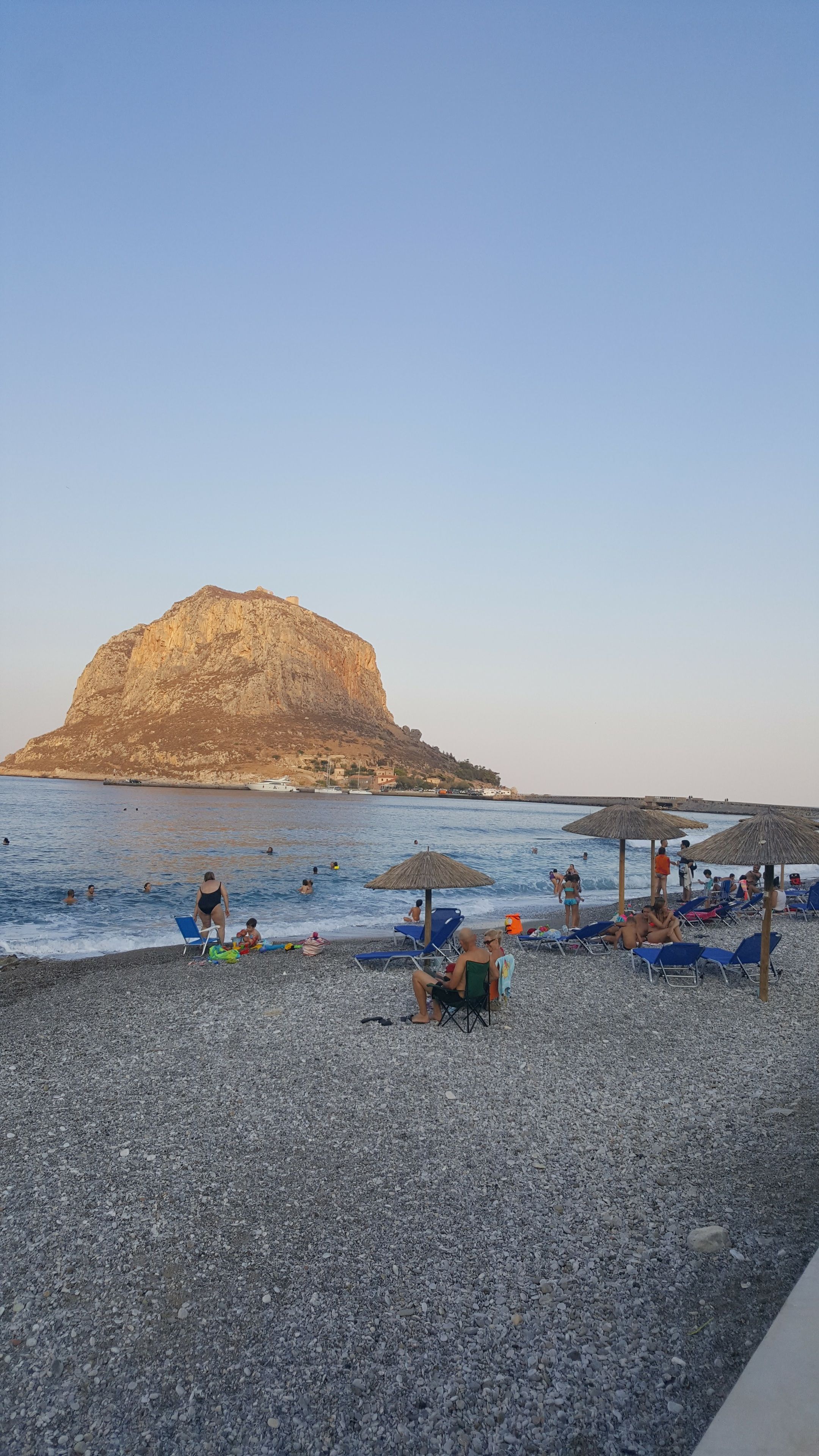 Una playa de guijarros con sombrillas y sillas, una gran formación rocosa al fondo y gente nadando y relajándose.