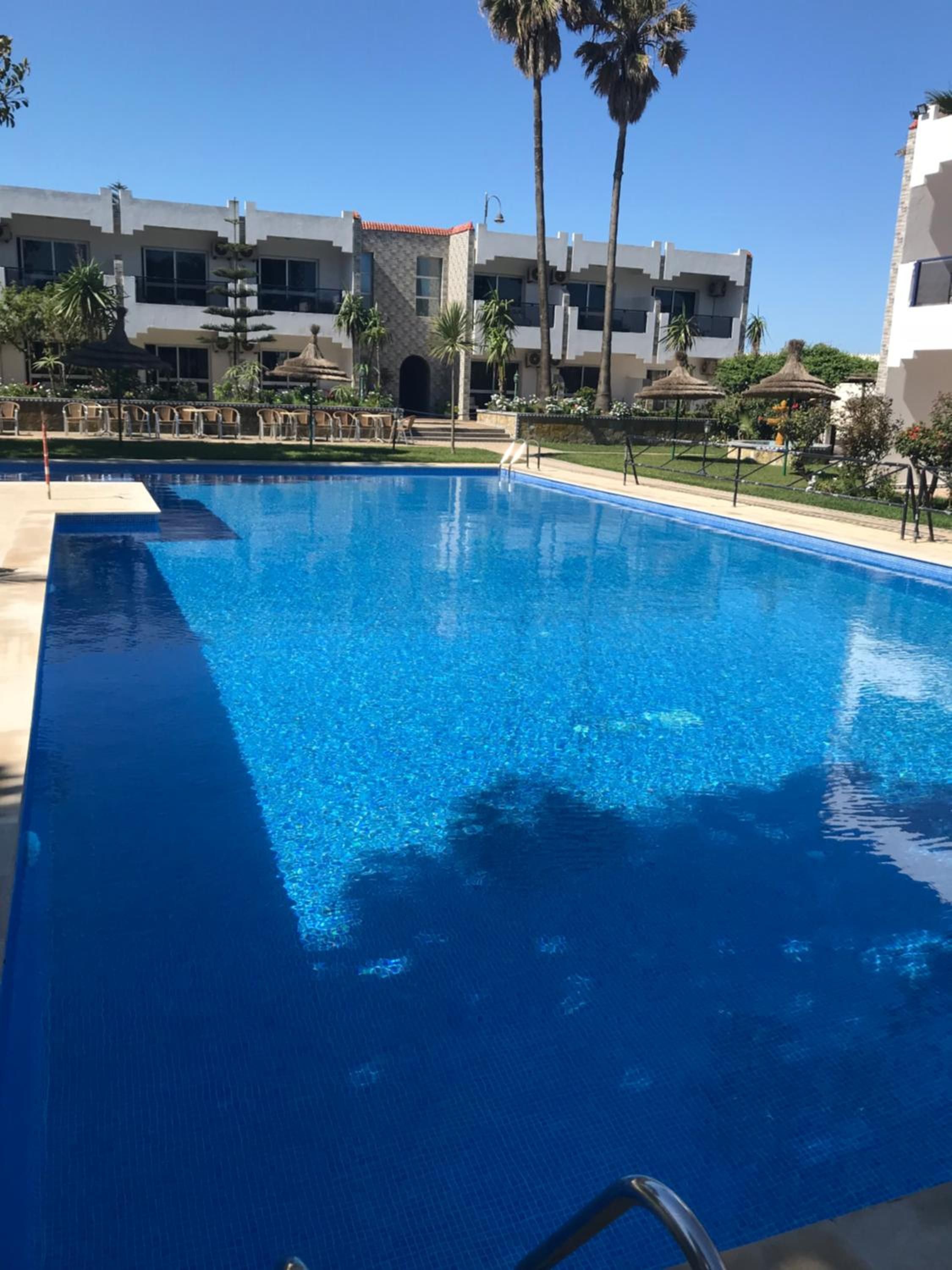 Una piscina azul cristalina con zona de borde de piscina, palmeras y un edificio con estilo arquitectónico moderno al fondo.