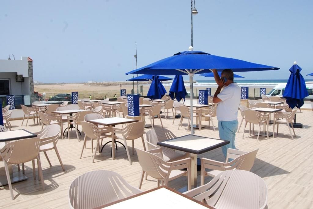 An outdoor seating area with tables and chairs, some with blue umbrellas, and a view of the beach.