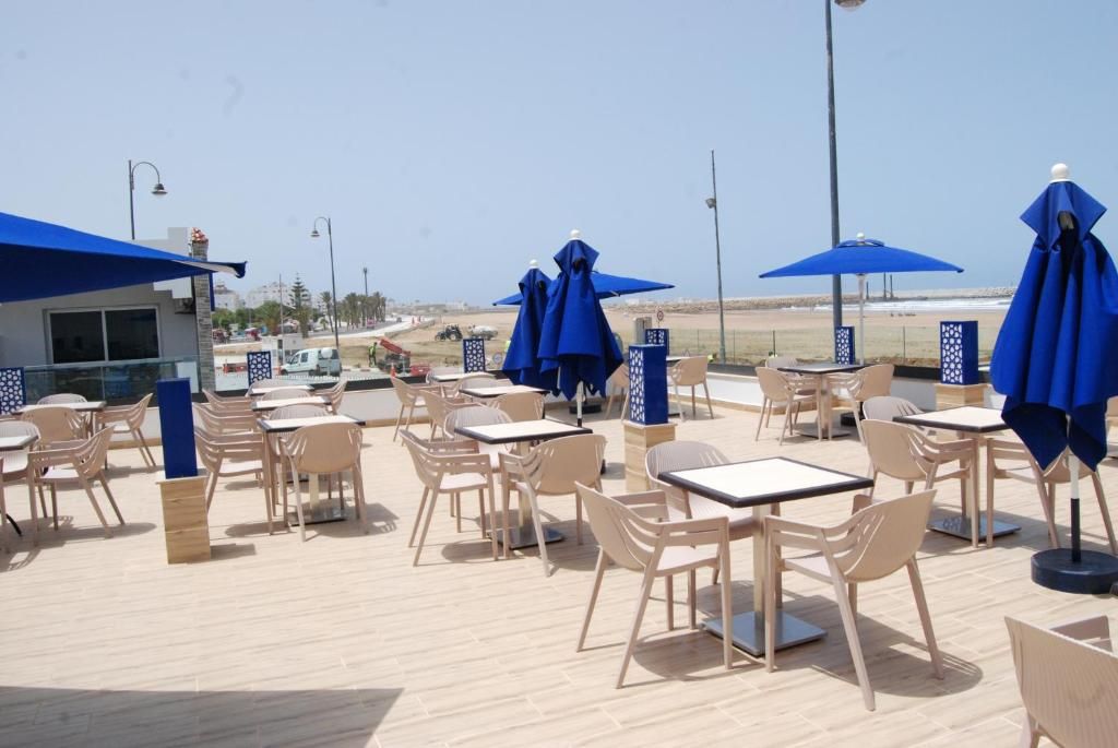 Un área de estar al aire libre con mesas y sillas, sombrillas azules y vista a la playa.