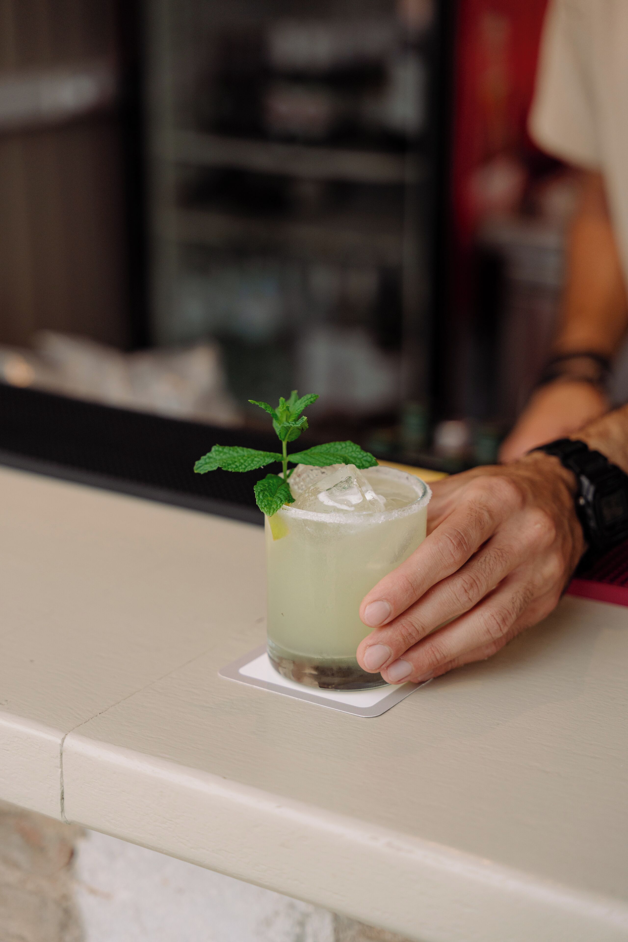 Une main tenant un verre décoré d’une feuille de menthe, posé sur une surface blanche.