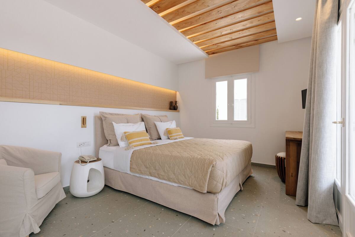 A bedroom with a bed, a bedside table, a chair, a window, and a wooden ceiling.