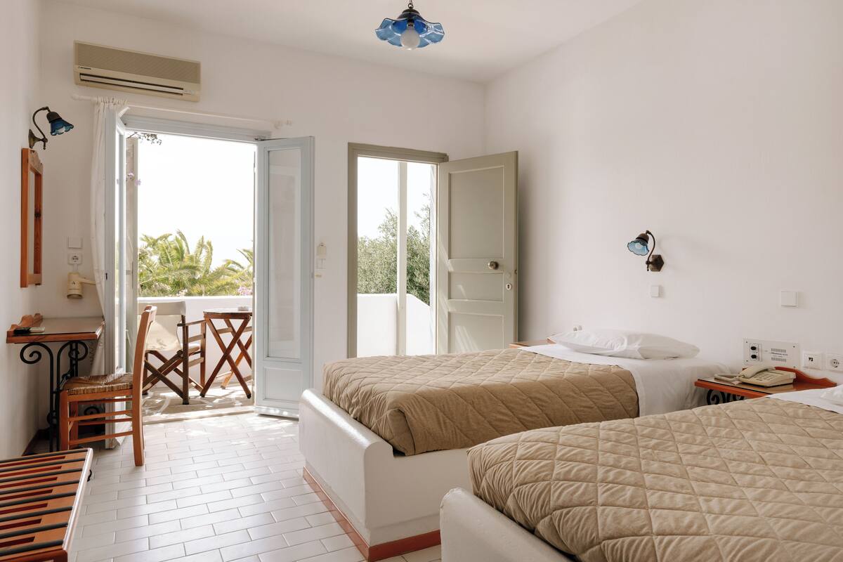 A hotel room with two beds, a desk, a chair, and a balcony with a view of palm trees.