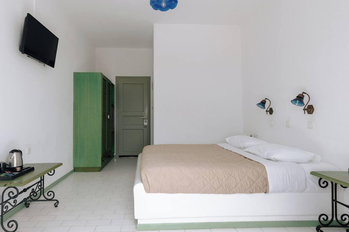 A bedroom with a bed, a TV mounted on the wall, a table with a kettle, and a green door.
