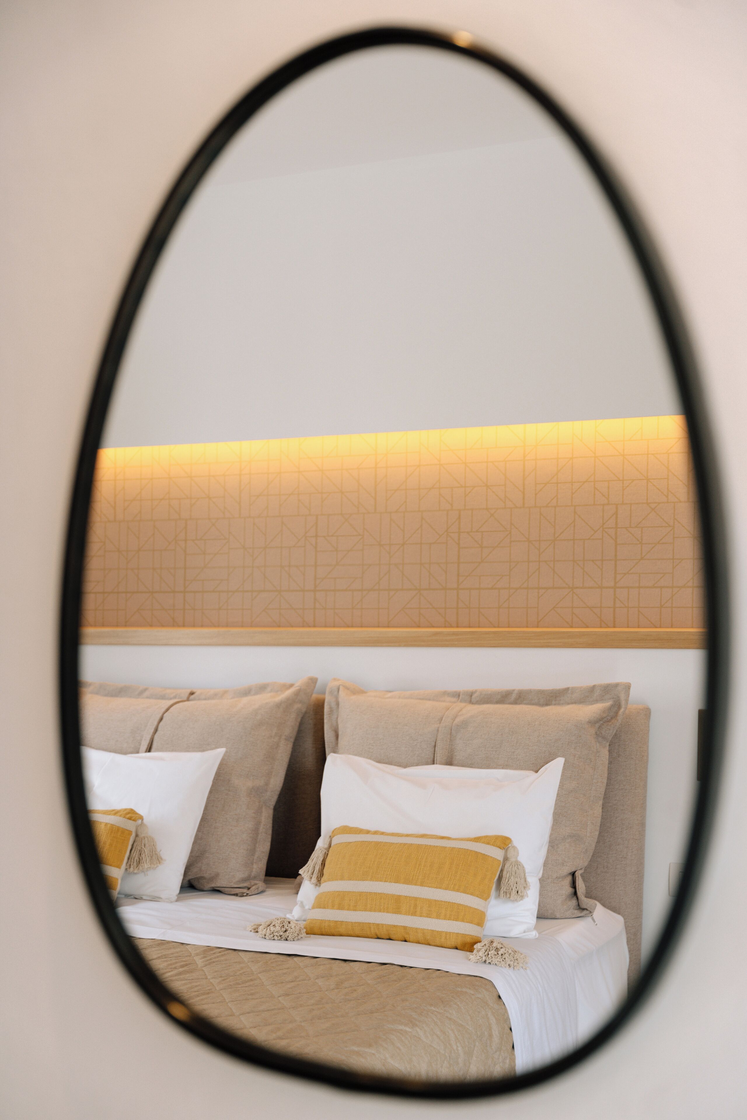 A bed with beige bedding and pillows, a headboard with a geometric pattern, and a mirror reflecting the room.