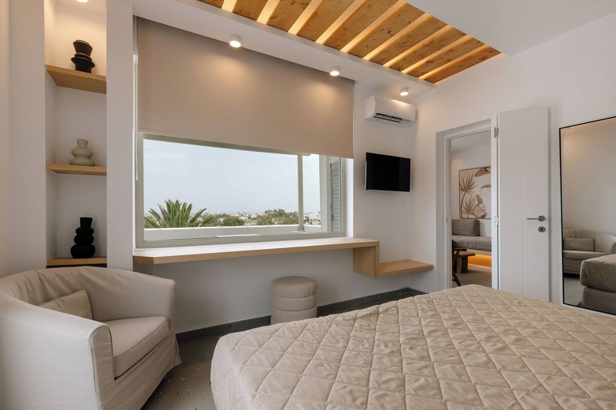 A modern bedroom with a large bed, a window with a view, built-in shelves, and a flat-screen TV.