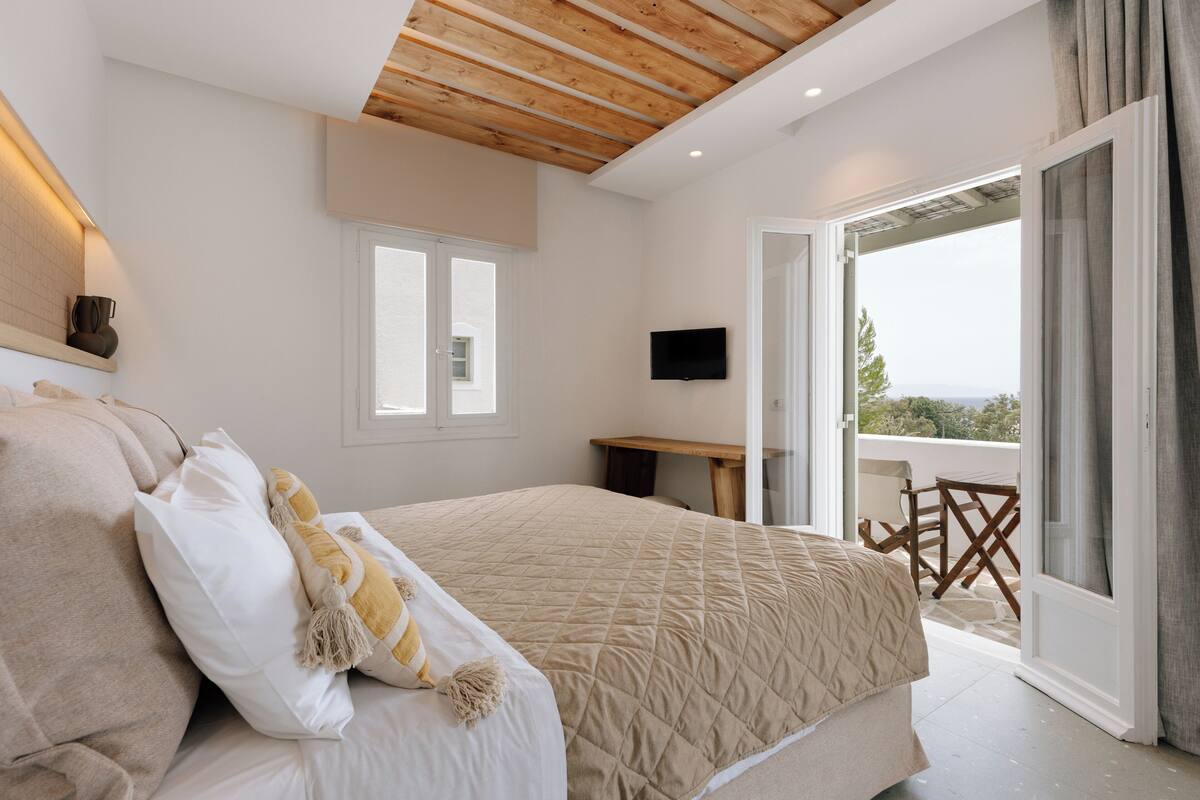 A bedroom with a bed, a TV, a balcony with a table and chairs, and a view of the outdoors.