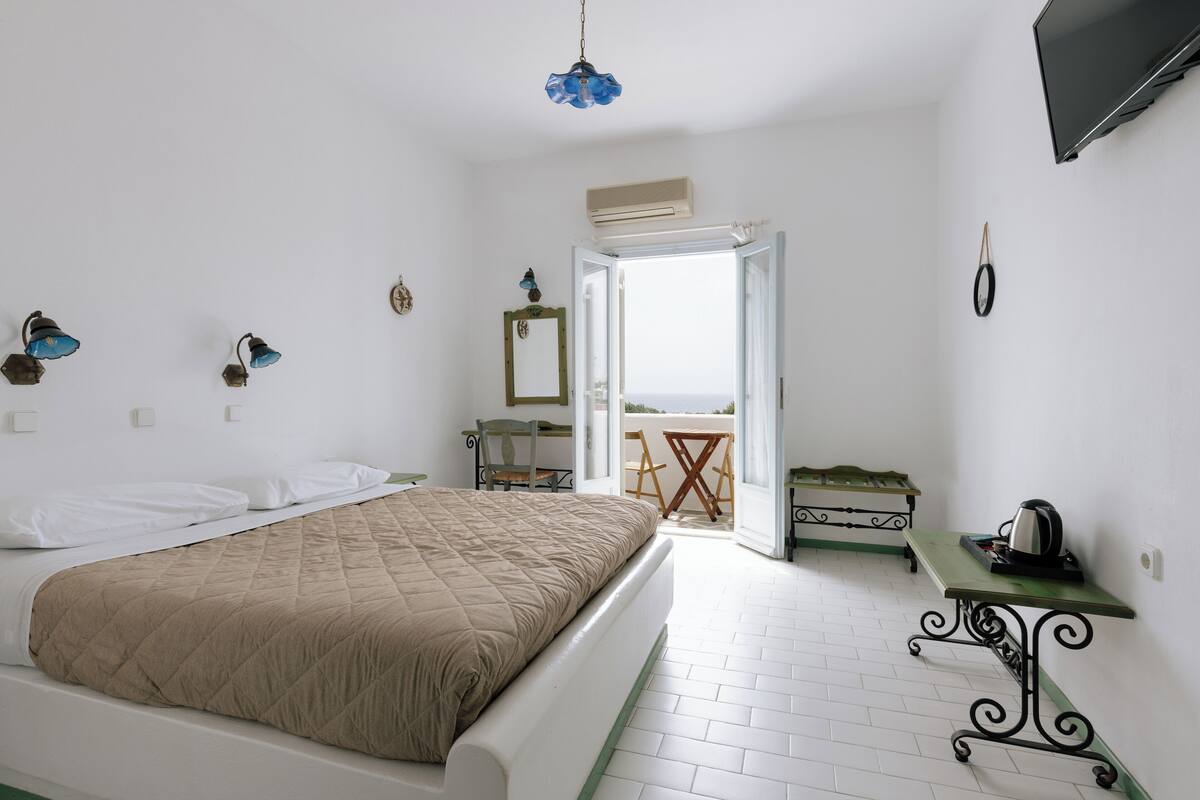 A bedroom with a bed, a table with a kettle, a chair, and a balcony with a view.