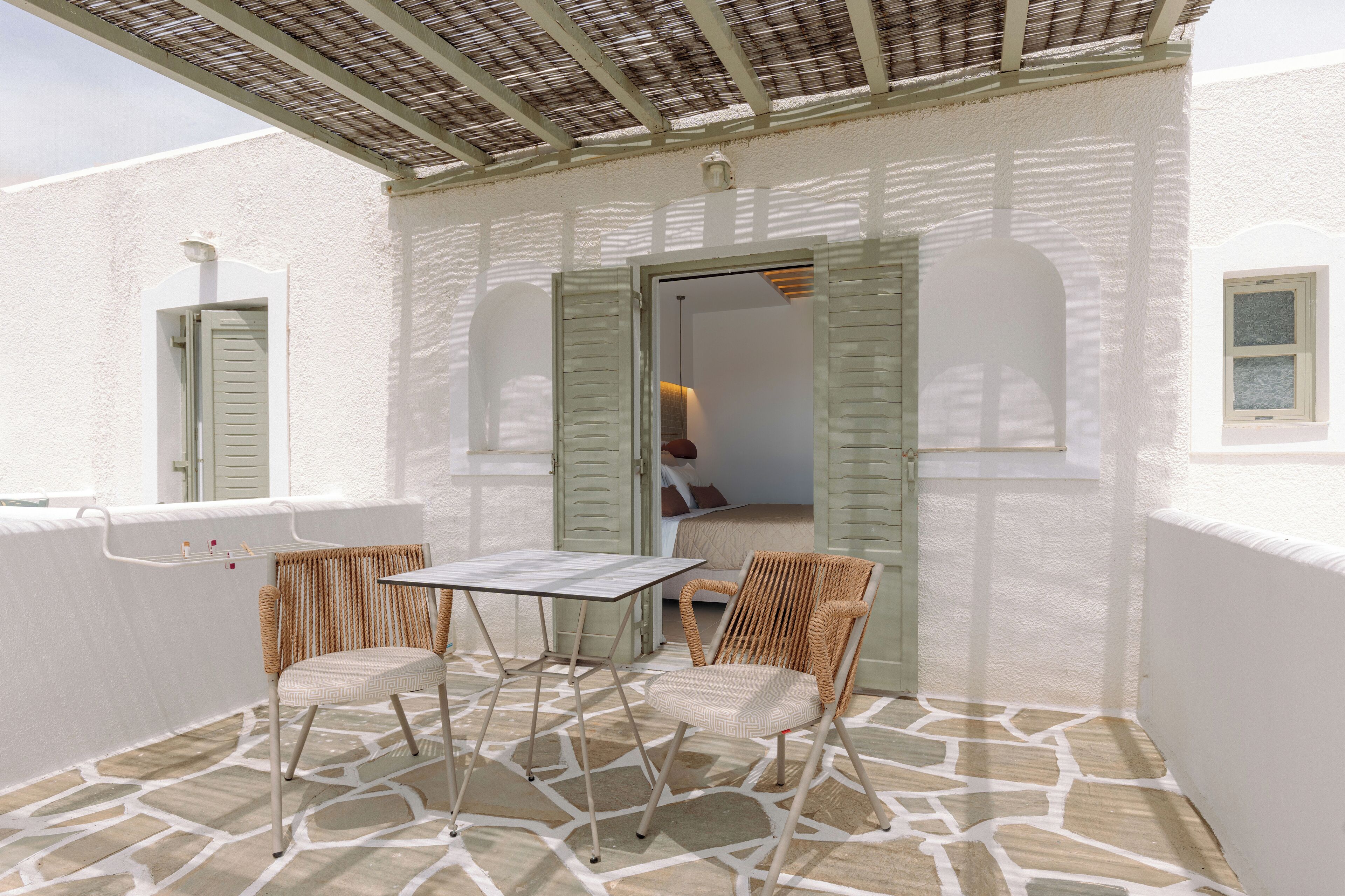 Eine Terrasse im Freien mit Tisch und zwei Korbstühlen, eine weiße Wand mit Fenster sowie eine Tür zum Innenbereich.