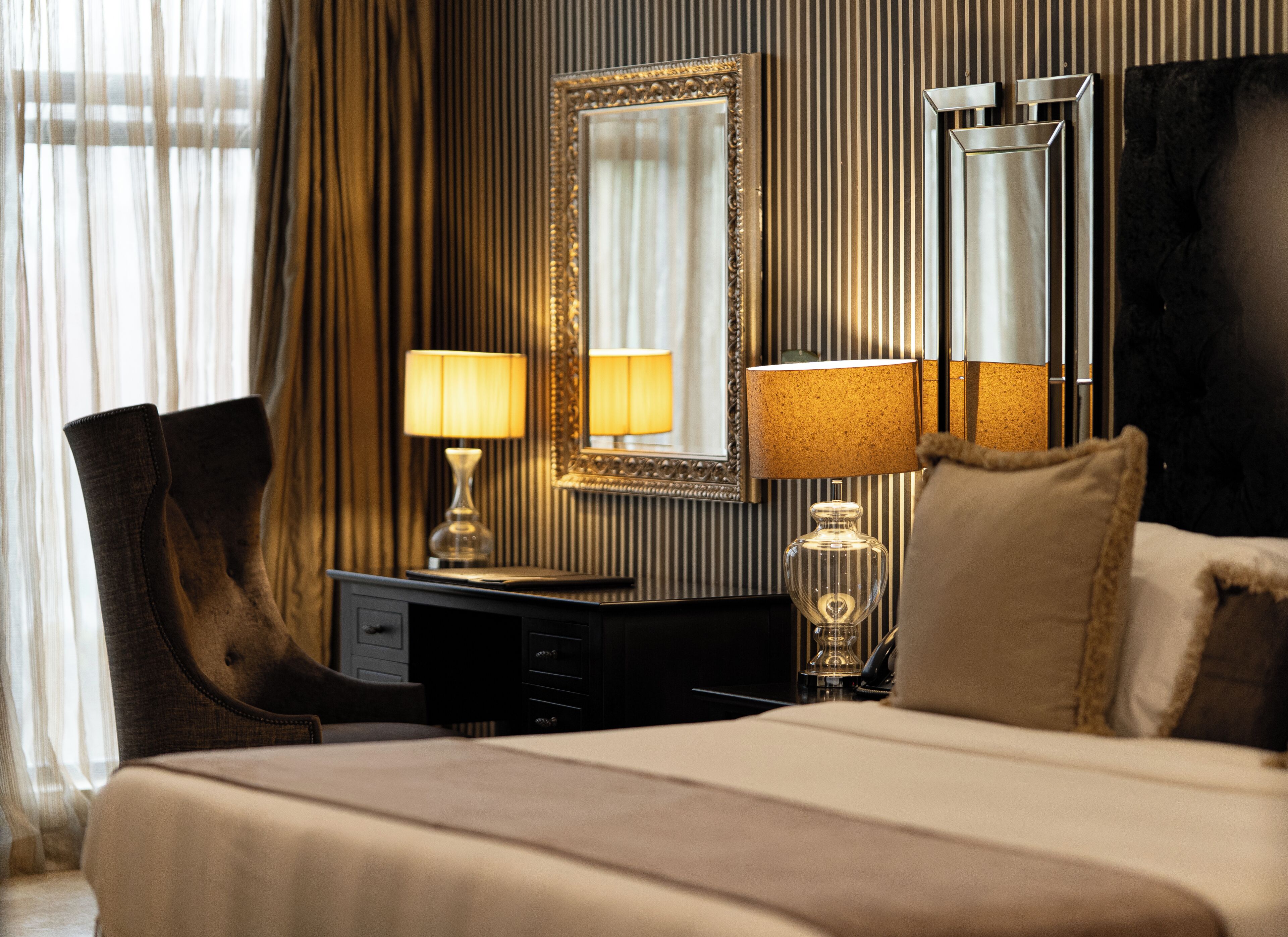 Habitación de hotel con cama, dos lámparas de noche, un escritorio con lámpara, una silla, un espejo grande y una ventana con cortinas.