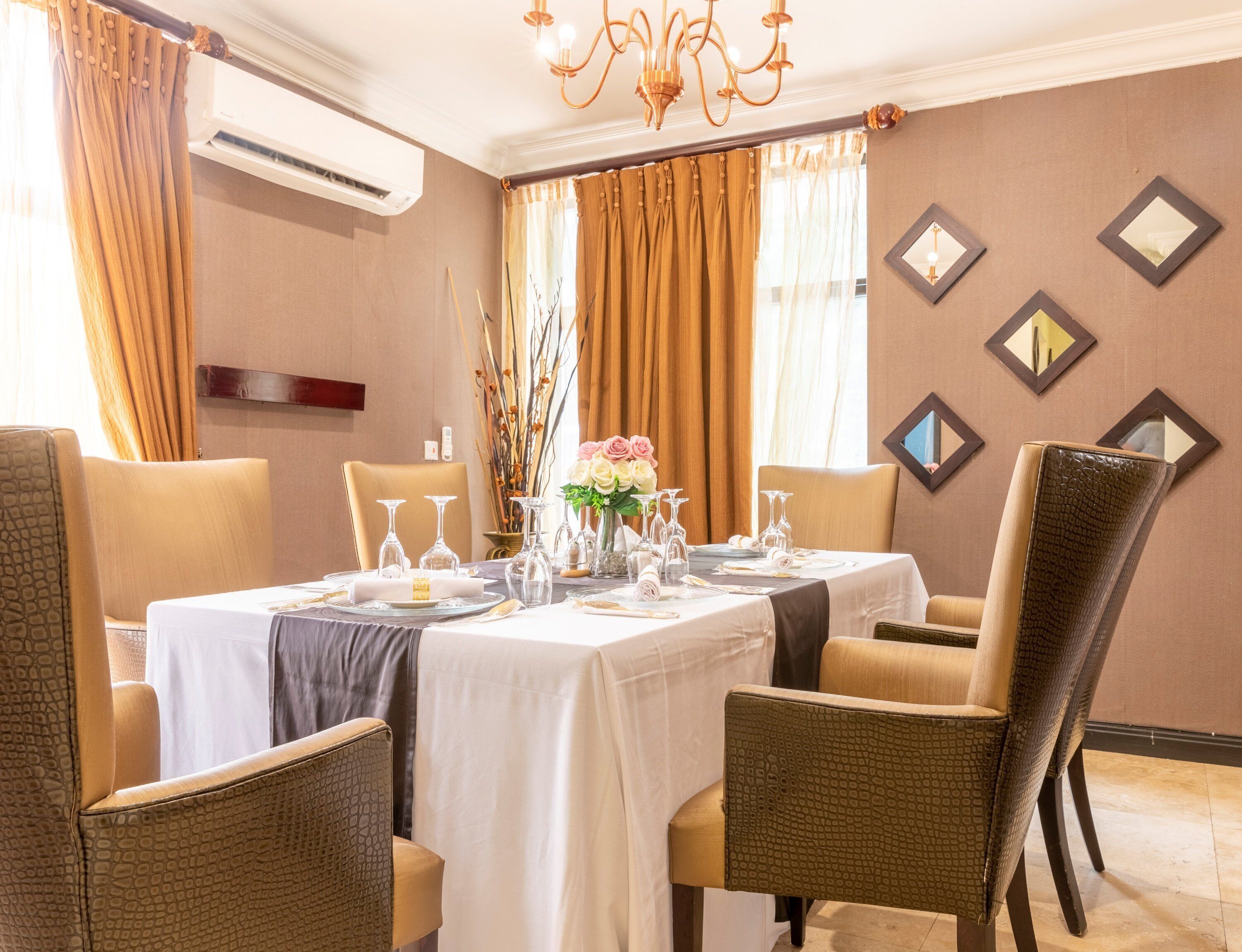 A dining area with a table set for four, surrounded by chairs, with a chandelier above and a wall-mounted air conditioner to the left.