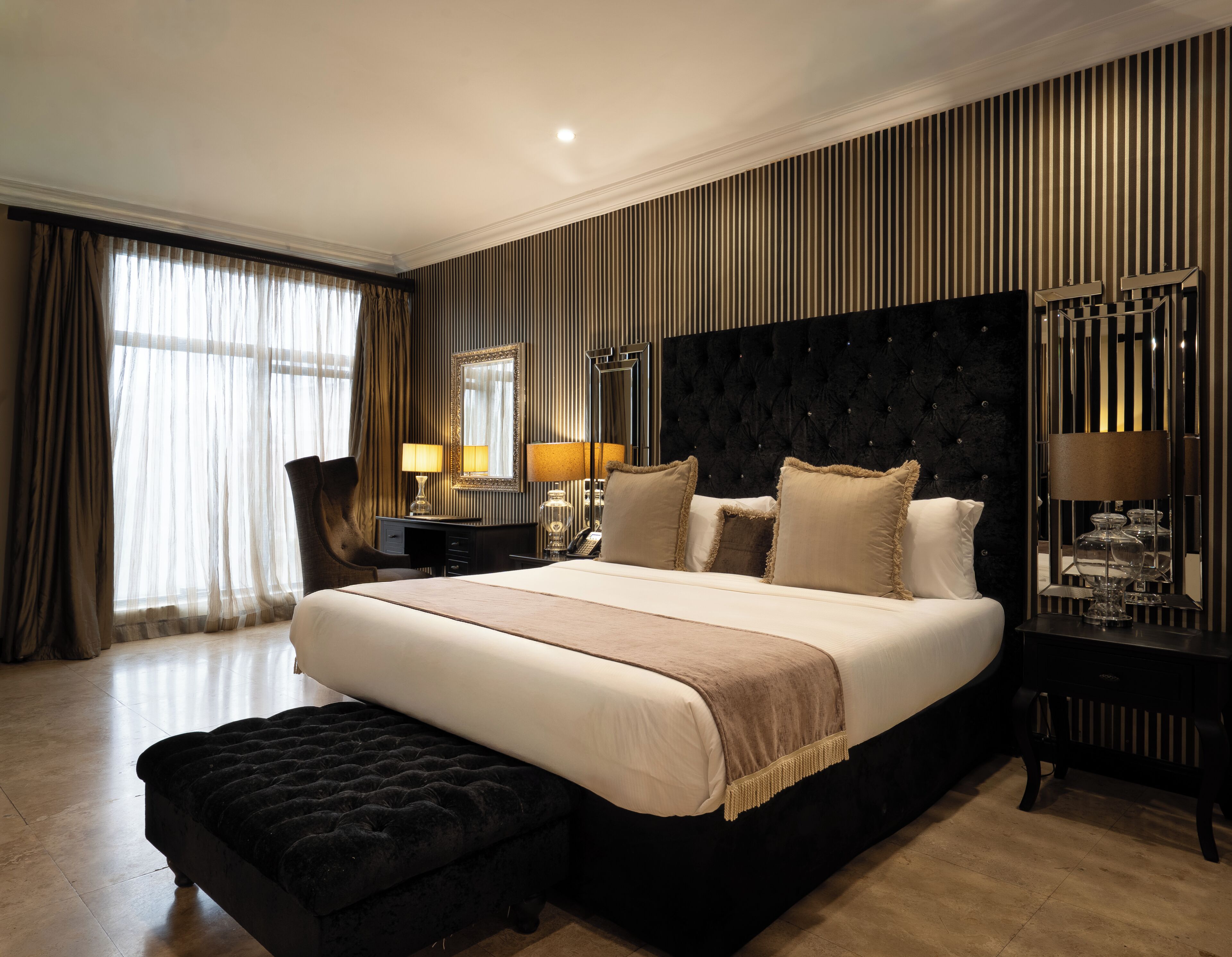 Ein Hotelzimmer mit einem großen Bett, einem Schreibtisch, einem Stuhl, einem Nachttisch mit Lampen und einem großen Spiegel.