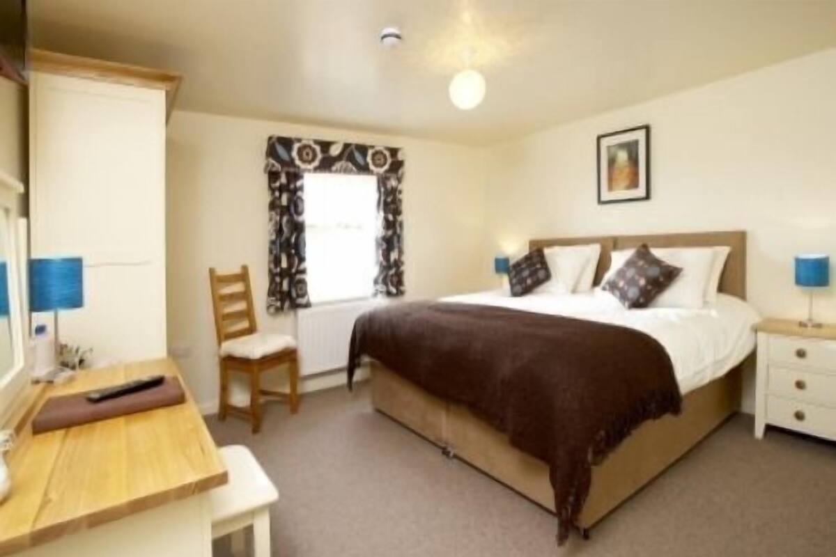 A bedroom with a bed, a desk, a chair, a nightstand, and a window with curtains.
