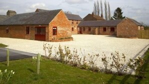 Un bâtiment en pierre avec une porte rouge, une allée gravillonnée et une zone herbeuse plantée.