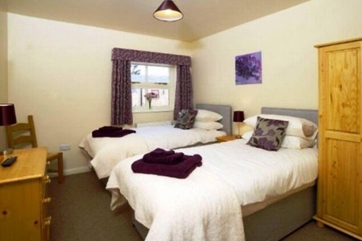 A hotel room with two beds, a wooden wardrobe, a desk with a lamp, and a window with curtains.
