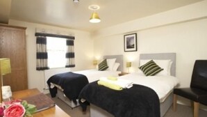 Une chambre d’hôtel avec deux lits, un bureau, une chaise, une fenêtre avec des rideaux, un tableau au mur et un vase avec des fleurs.
